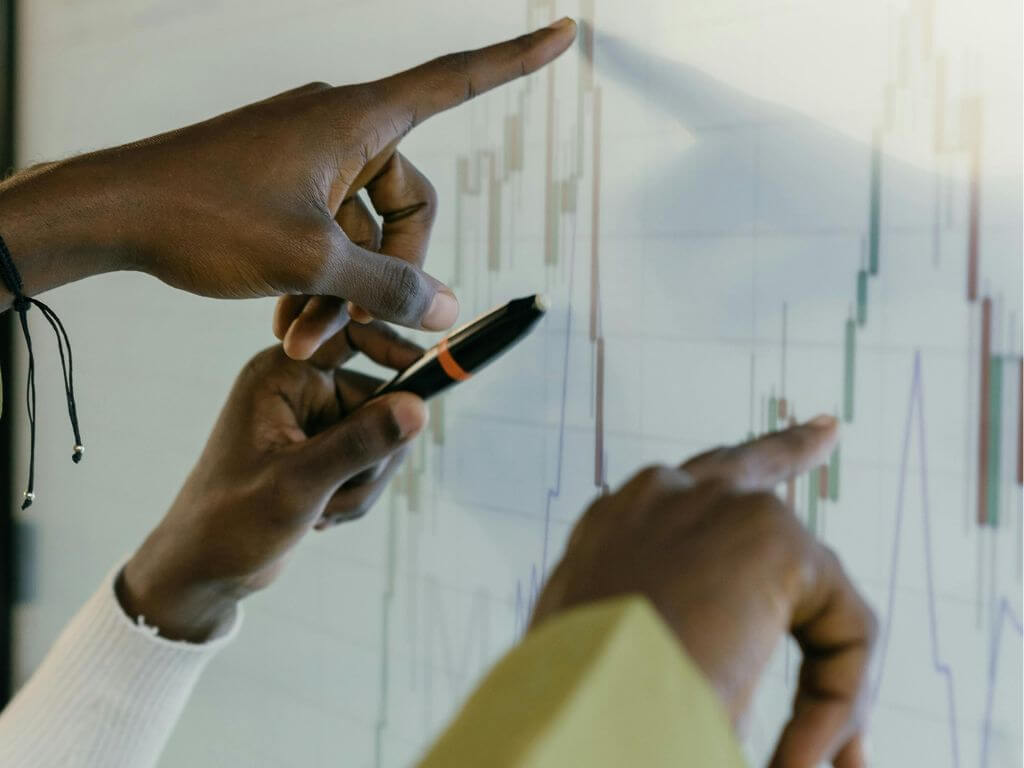 Close-up of two hands, one holding a smartphone and the other pointing at a transparent board with financial graphs.