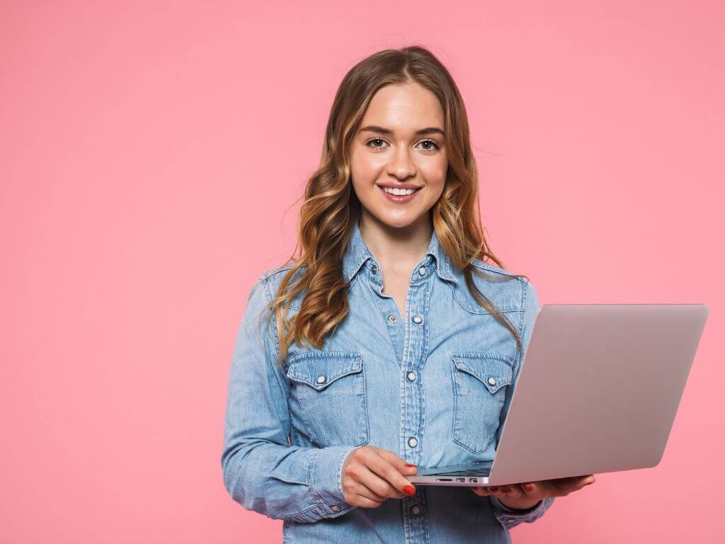 Smiling woman in a denim shirt holding an open laptop against a pink background.