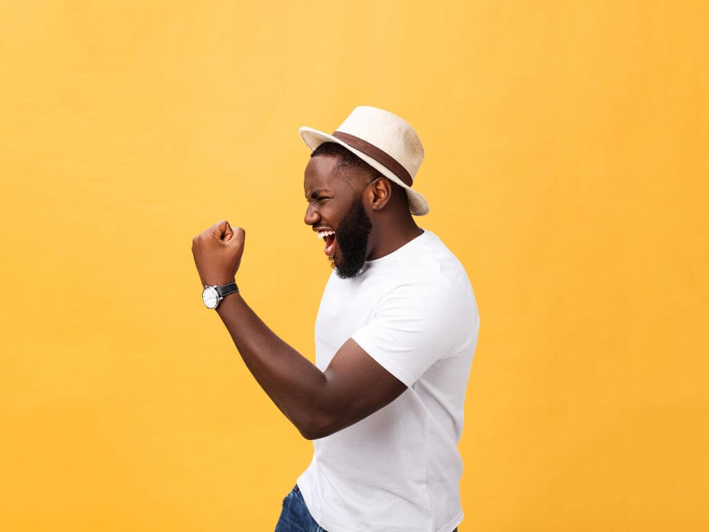 Joyful man in a white t-shirt and hat punching the air in victory against a yellow background.