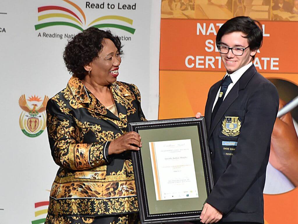 Basic Education Minister presenting a framed certificate to a proud male student on stage.