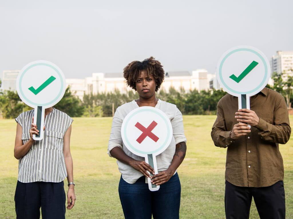 Three people standing in a field, each holding a sign. Two signs show green checkmarks, and the middle sign shows a red X.