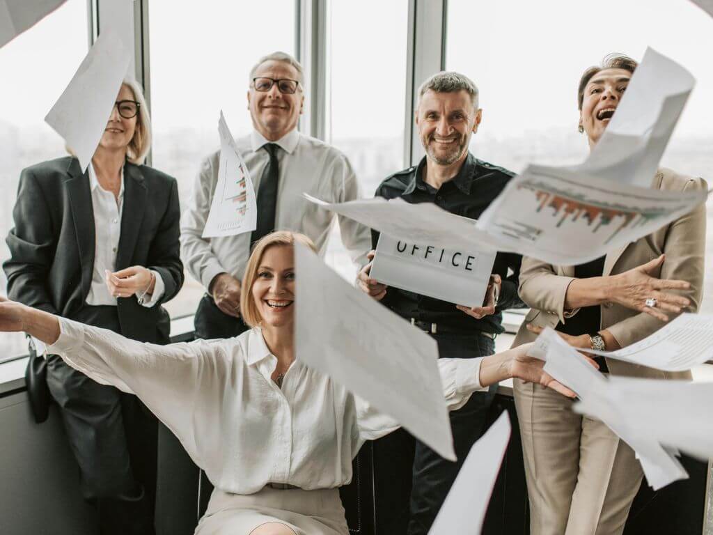 Group of business professionals celebrating in an office, throwing papers in the air with smiles on their faces.