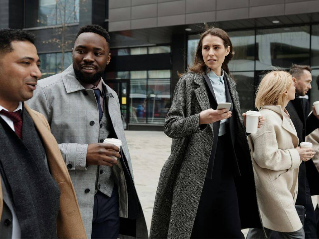 Group of business professionals walking outside an office building, holding coffee cups and engaged in conversation.