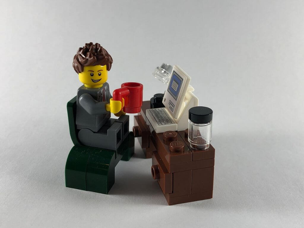 A playful LEGO figure seated at a desk with a laptop and coffee cup