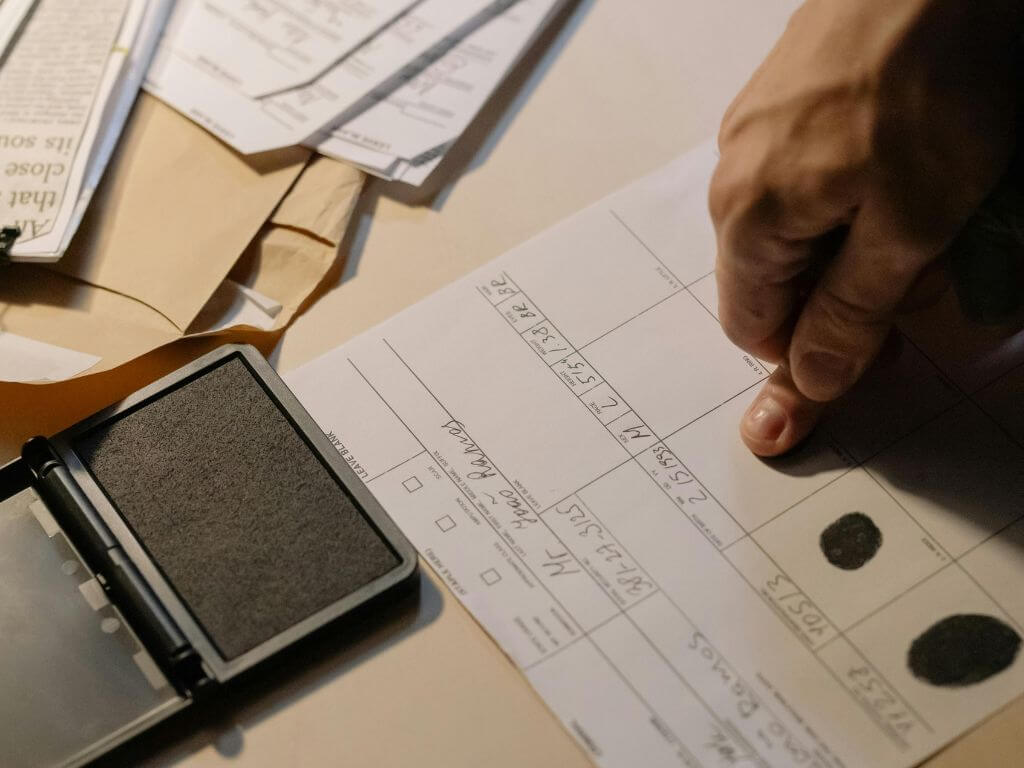 Close-up of a person applying fingerprints to a police record form, with an ink pad on a desk surrounded by paperwork